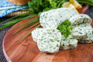 Slices of Kräuterbutter (German Herb Butter) on a wooden board with chives and parsley in the background.