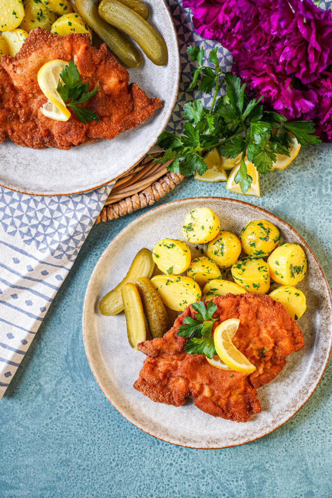 Aerial view of Smažený Vepřový Řízek (Czech Breaded Pork Cutlet) on two plates with parsley potatoes and pickles.