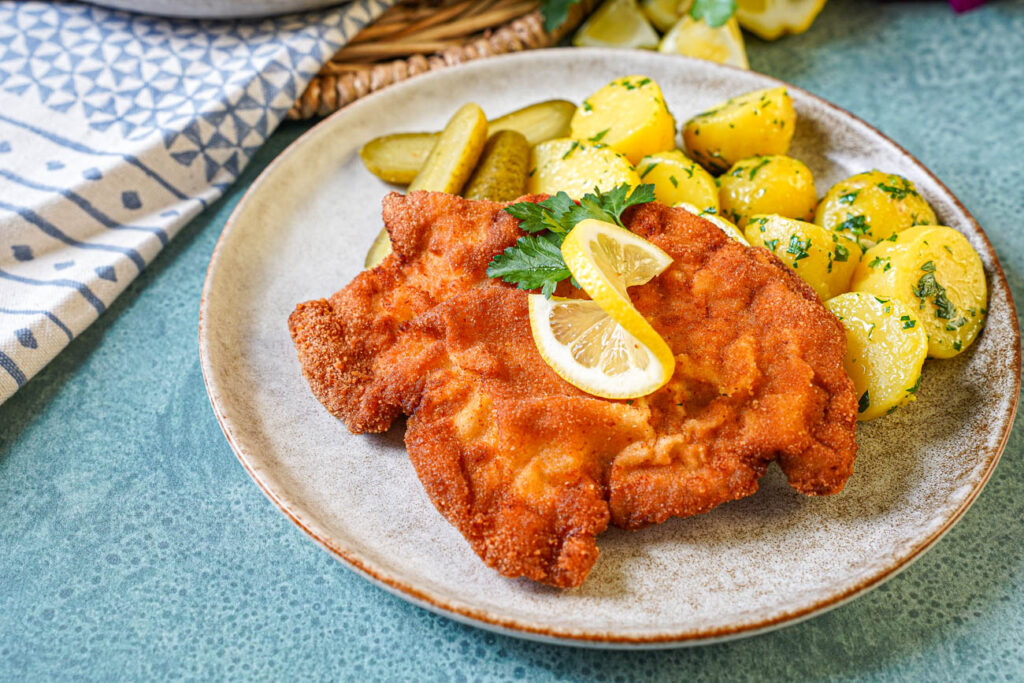 Smažený Vepřový Řízek (Czech Breaded Pork Cutlet) on a plate with a lemon slice, pickles, and parsley potatoes.