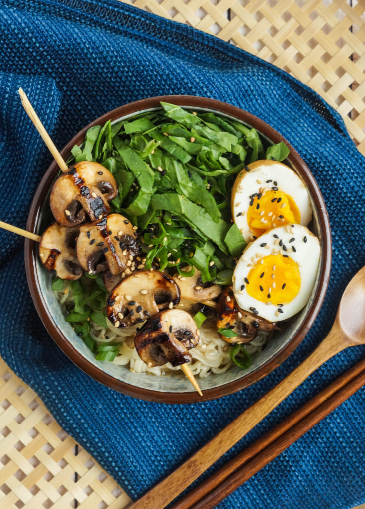 Aerial view of Grilled Mushroom Noodle Bowls in a bowl with noodles, grilled mushrooms on two skewers, shredded spinach, and two halves of a hard boiled egg next to a wooden spoon and chopsticks.
