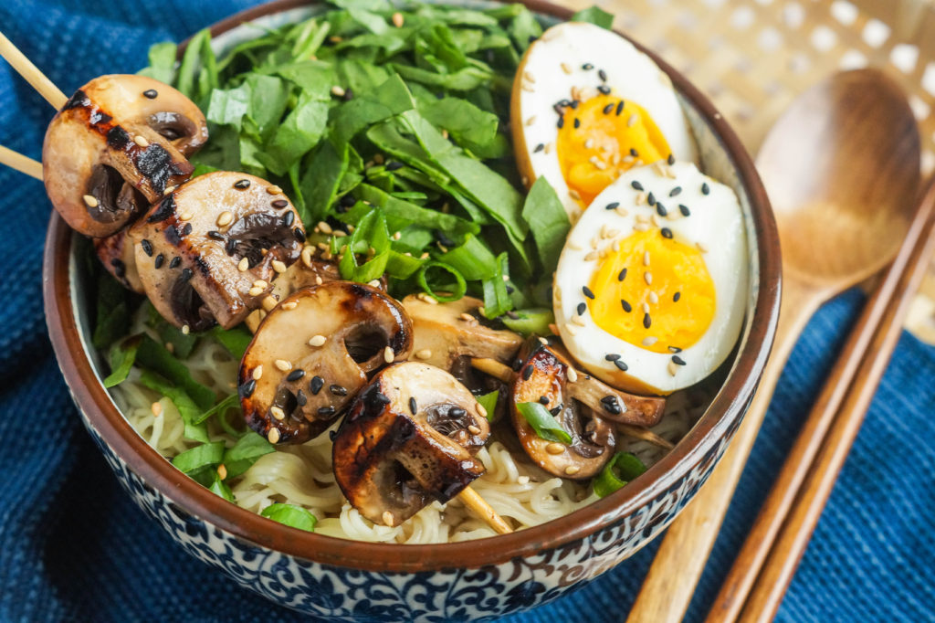 Grilled Mushroom Noodle Bowls topped with two skewers of grilled mushrooms, shredded spinach, and 2 halves of a hardboiled egg over a bed of noodles.