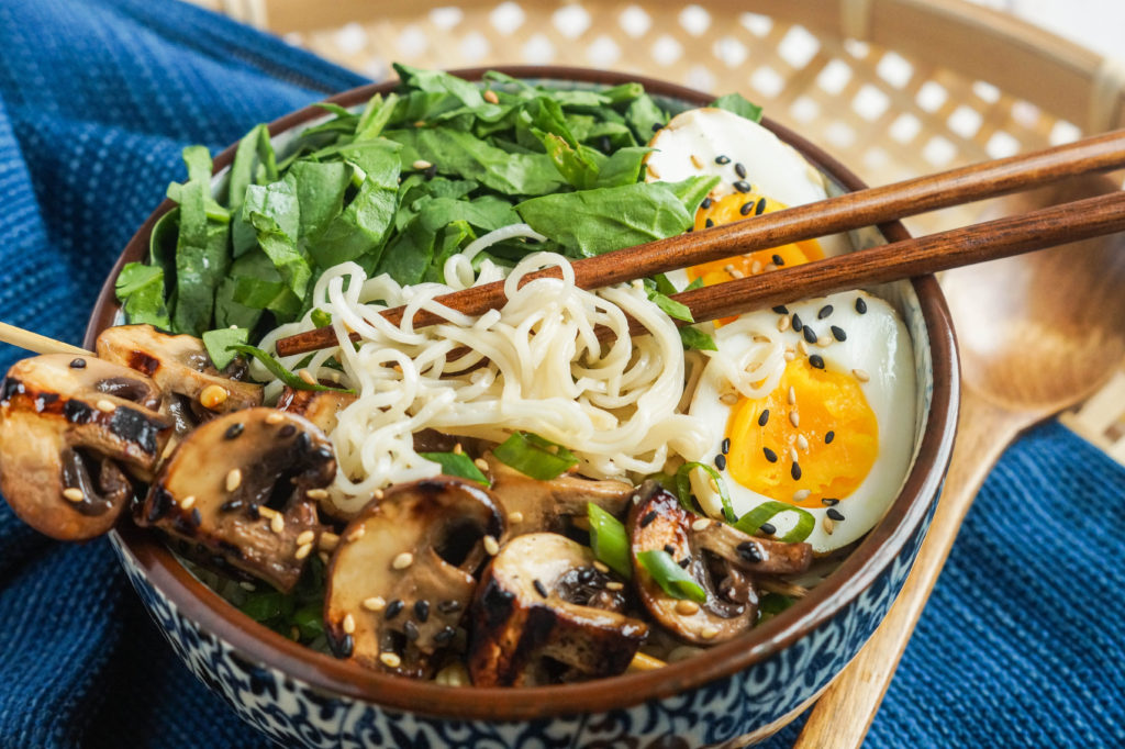 Chopsticks lifting noodles from the Grilled Mushroom Noodle Bowls with grilled mushrooms, shredded spinach, and two halves of a hard-boiled egg.