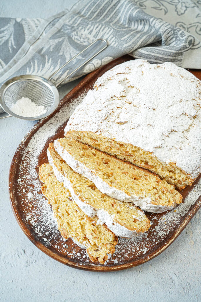 Aerial view of Quarkstollen (Quark-Almond Sweet Bread) on a wooden board next to a gray towel and powdered sugar in a sieve.