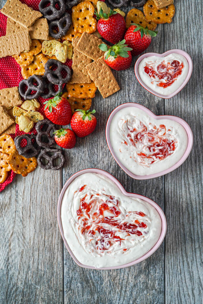 Aerial view of Strawberry Cheesecake Dip in three heart shaped bowls next to pretzels, graham crackers, and strawberries.