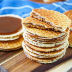 A stack of Stroopwafels (Dutch Syrup-Filled Waffle Cookies) on a wooden board next to a blue towel.
