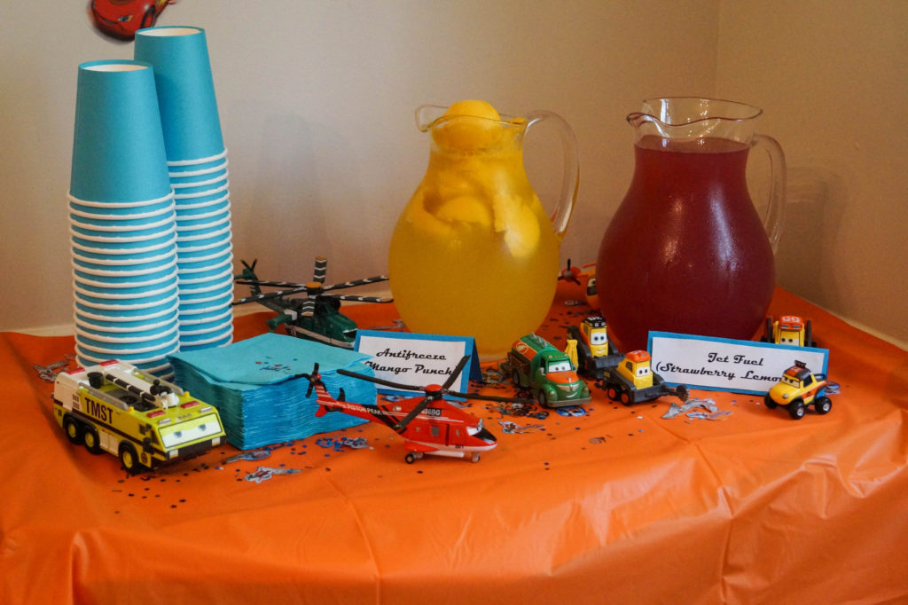 Cars and Planes drink table with two glass pitchers, paper cups, and cars/planes toys.