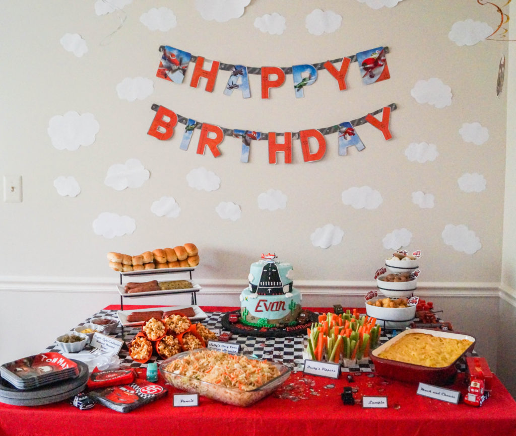 Red table covered in food, and cake, and a happy birthday banner on the wall.
