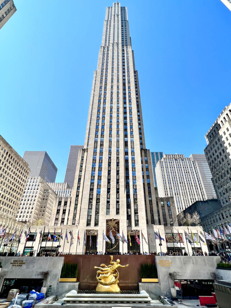 View of Rockefeller Center with flags and golden statue in front of tall building.