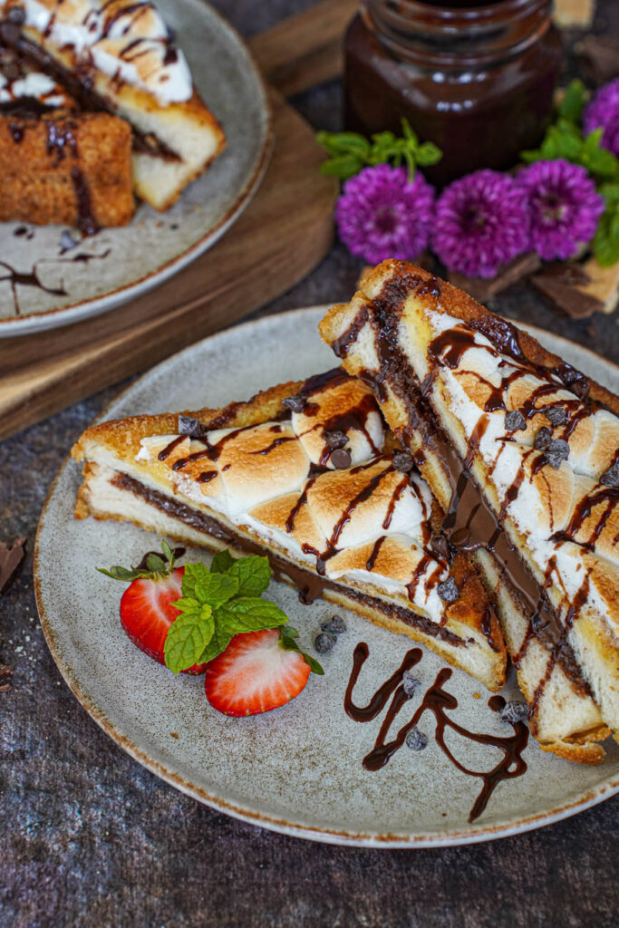 Close up of S'mores French Toast on two plates with purple flowers and a jar of chocolate sauce in the background.