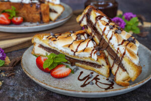 Two S'mores French Toast halves on a plate with strawberries, mint sprigs, and chocolate sauce.