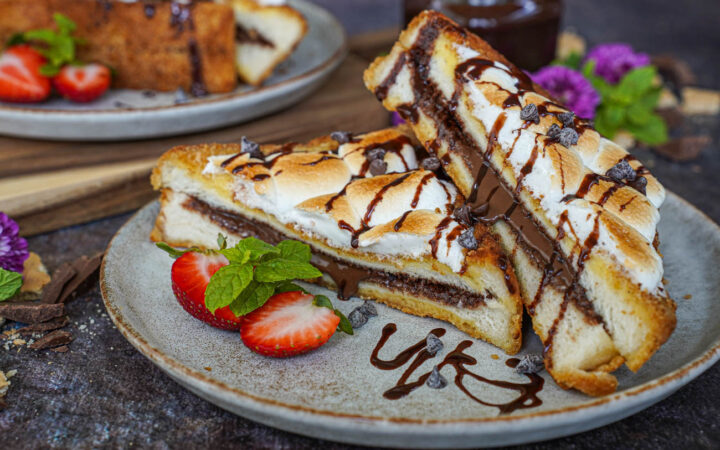Two S'mores French Toast halves on a plate with strawberries, mint sprigs, and chocolate sauce.