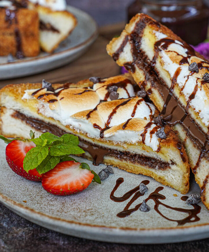 Two S'mores French Toast halves on a plate with strawberries, mint sprigs, and chocolate sauce.