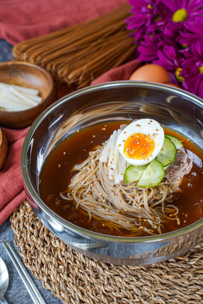 Close up of Mul Naengmyeon (Buckwheat Noodles in Chilled Broth) in a metal bowl with eggs, purple flowers, buckwheat noodles, and pear in the background.