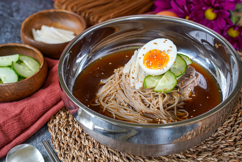 Mul Naengmyeon (Buckwheat Noodles in Chilled Broth) in a metal bowl next to wooden bowls of cucumber and pear.
