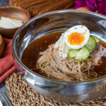 Mul Naengmyeon in a metal bowl next to wooden bowls of cucumber and pear slices.