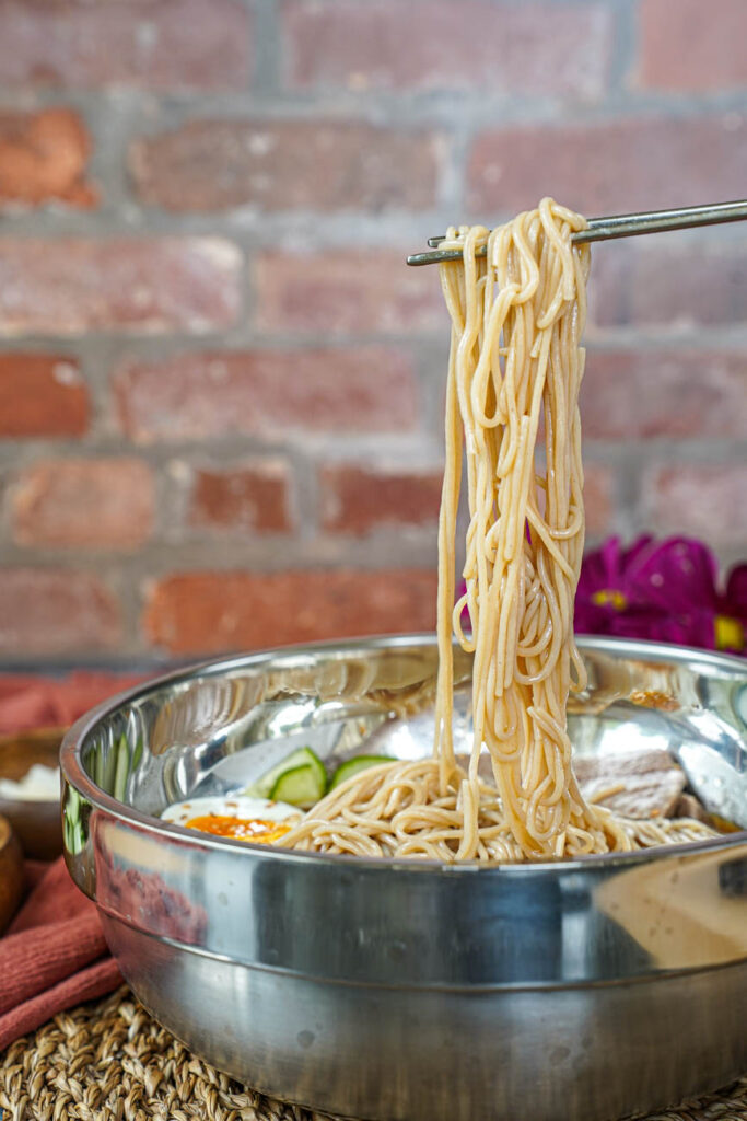 Chopsticks holding buckwheat noodles over a metal bowl.
