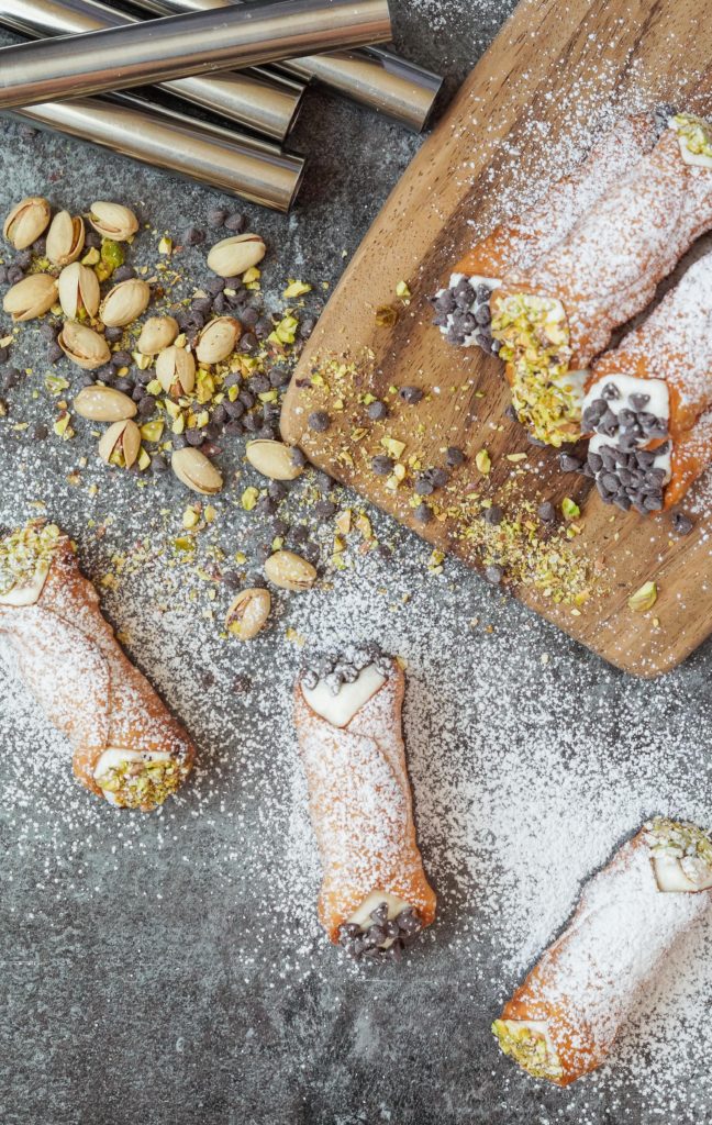 Aerial view of Cannoli topped with powdered sugar and next to scattered pistachios and mini chocolate chips.