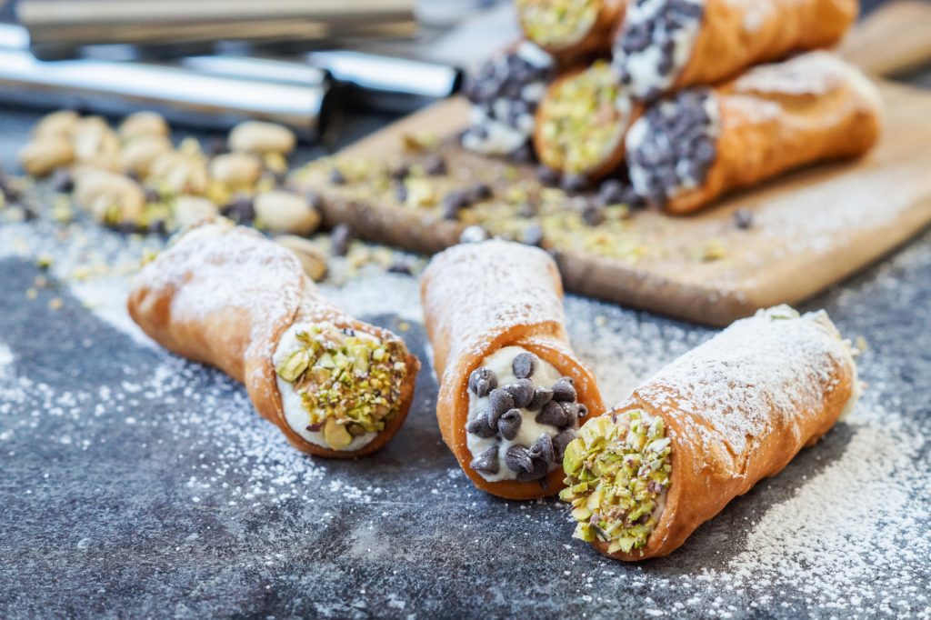 Three cannoli covered in powdered sugar with more stacked in the background on a wooden board.