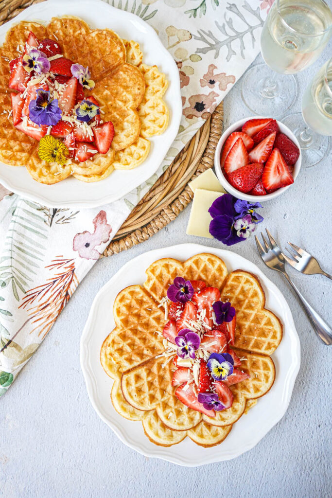 Aerial view of Prosecco Waffles on two plates next to flowers, white chocolate, strawberries, and wine glasses.