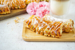 Two Nusskämme (Hazelnut Combs) on a wooden board with pink flowers and a latte macchiato in the background.