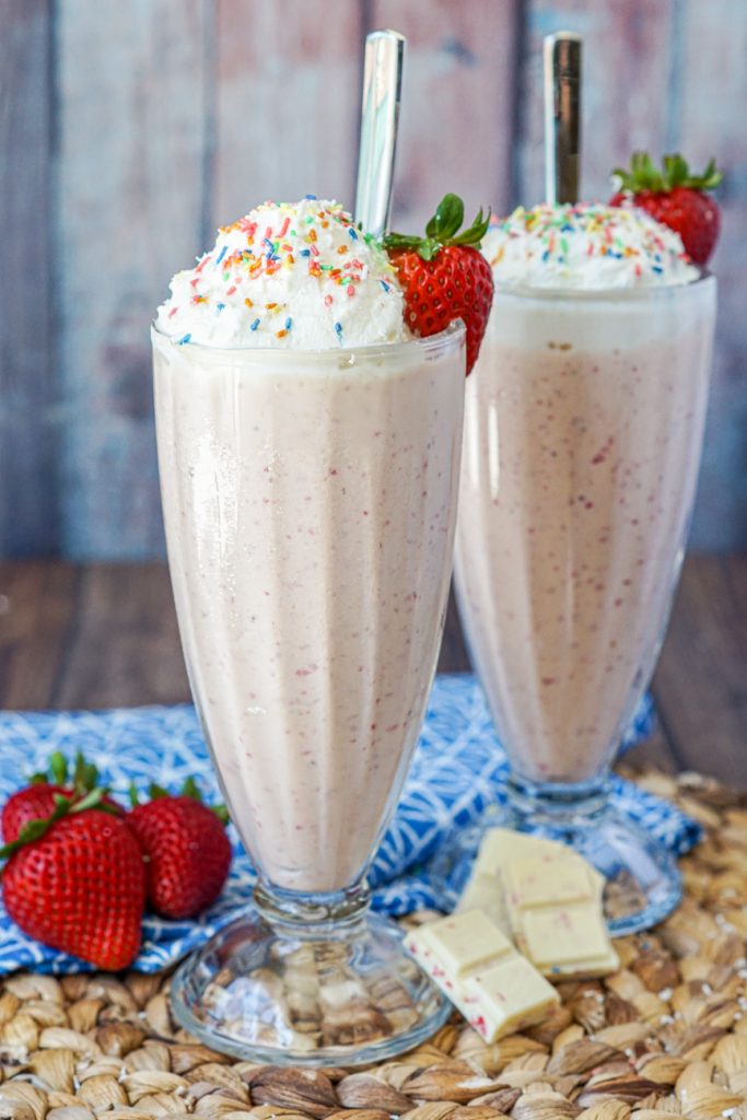 Strawberry White Chocolate Milkshake in two tall glasses with whipped cream, sprinkles, and metal spoons