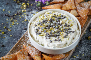 Cannoli Dip in a white bowl topped with mini chocolate chips and chopped pistachios and next to cannoli chips.