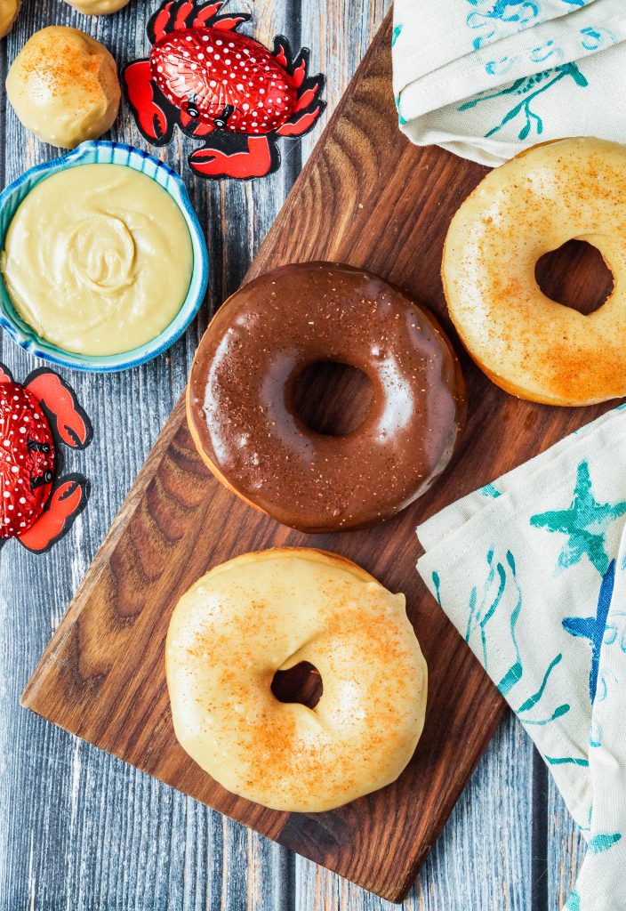 Aerial view of three Old Bay Doughnuts on a wooden board next to chocolate crabs and a bowl of caramel glaze.