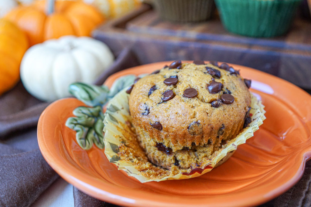 A muffin on a pumpkin plate with the wrapper pulled down and more Pumpkin Chocolate Chip Muffins in the background on a wooden board.