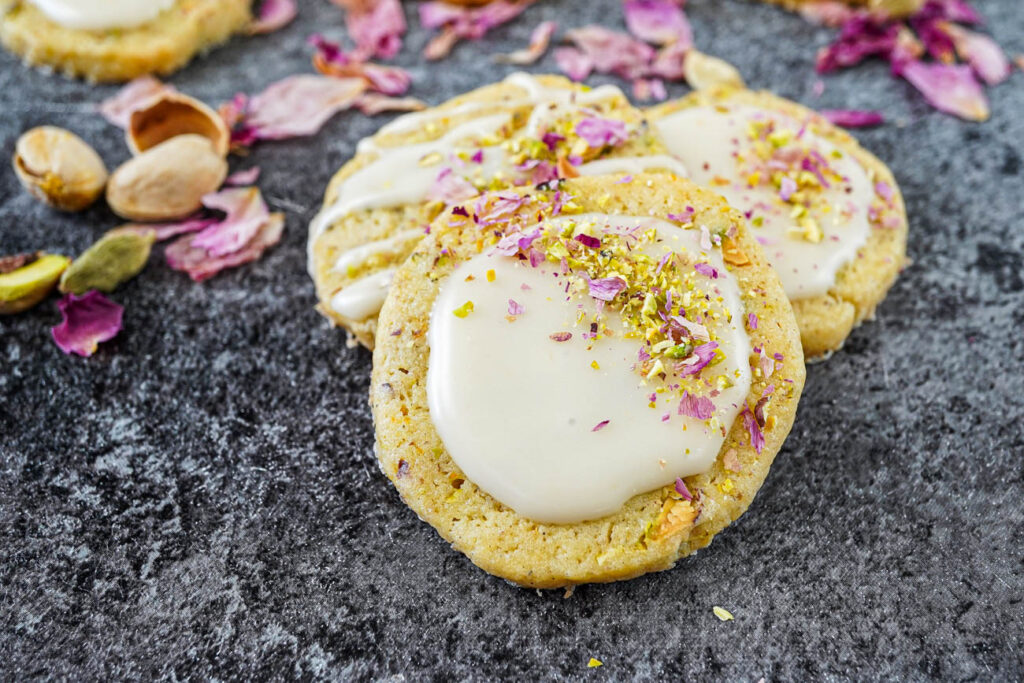 Three Orange Cardamom Cookies topped with a sweet glaze, chopped pistachios, and rose petals.