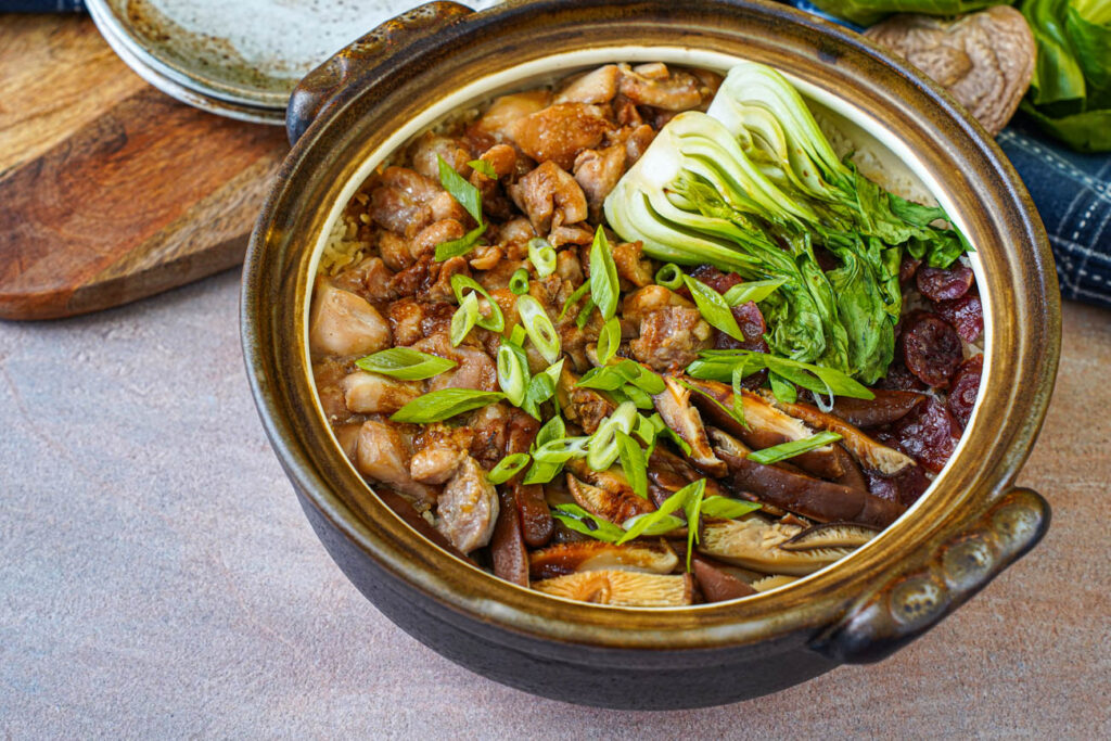 Clay Pot Chicken Rice in a brown clay pot with chicken pieces, sliced sausages, bok choy halves, and sliced mushrooms.