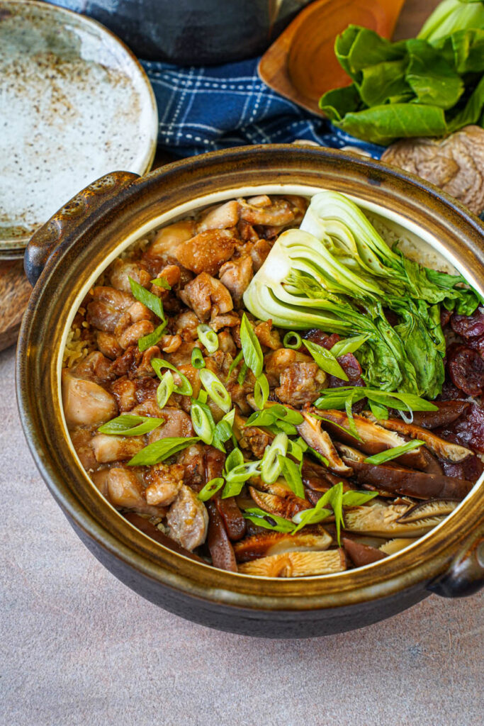 Clay Pot Chicken Rice in a brown pot, with plates, a wooden spoon, bok choy, and dried shiitake mushroom in the background.