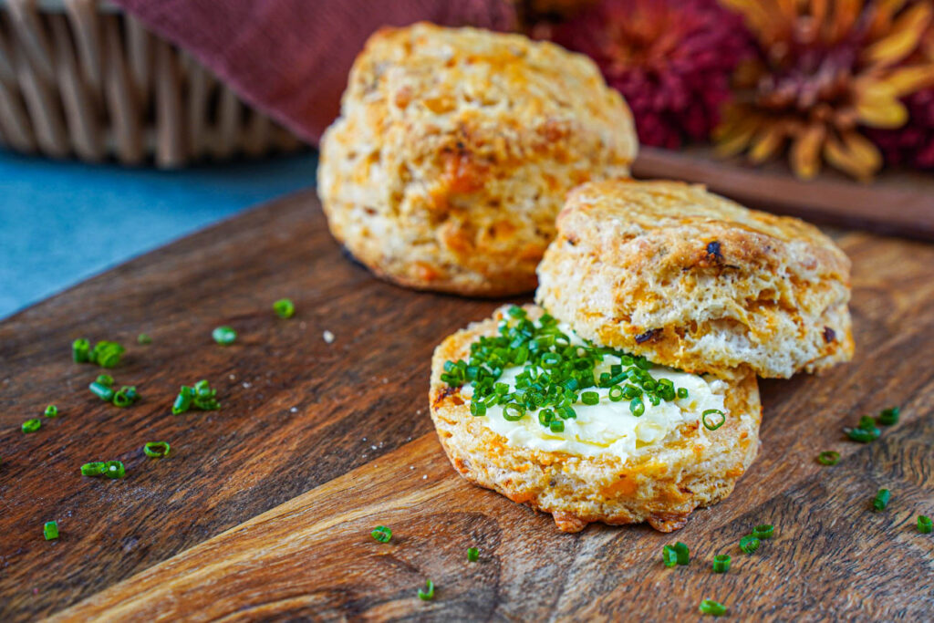 Two Cheddar Sun-Dried Tomato Biscuits on a wooden board with one cut in half and filled with butter and chopped chives.