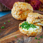 Two Cheddar Sun-Dried Tomato Biscuits on a wooden board with one cut in half and filled with butter and chopped chives.