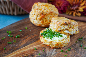 Two Cheddar Sun-Dried Tomato Biscuits on a wooden board with one cut in half and filled with butter and chopped chives.