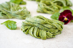 Three bundles of Homemade Spinach Fettuccine next to spinach leaves and a red pansy.