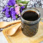 Homemade Teriyaki Sauce in a glass jar on a wooden board next to two wooden spoons and purple flowers.