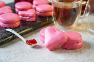 One heart shaped and two circle Napolitaines (Mauritian Sandwich Cookies) next to a cup of tea and spoonful of jam.