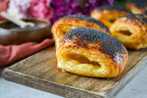 Tebirkes (Danish Poppyseed Pastries) on a wooden board with poppyseeds in a bowl and flowers in the background.