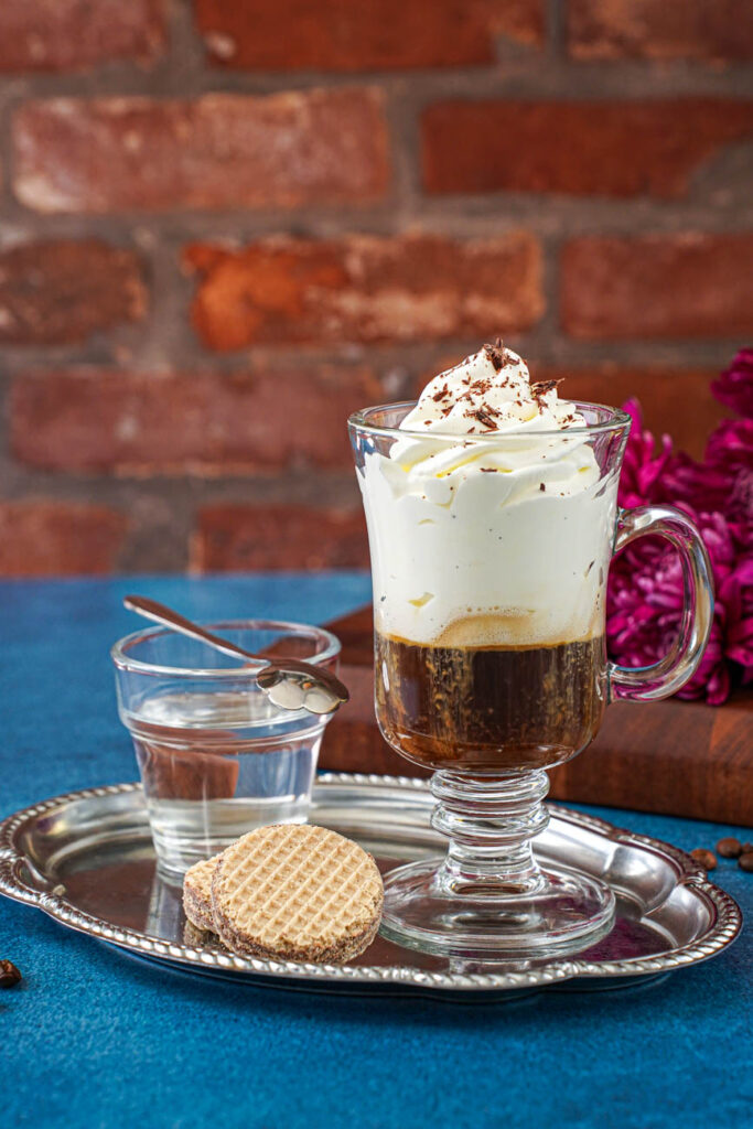 Side view of Einspänner (Viennese Coffee with Whipped Cream) on a tray with purple flowers in the background.