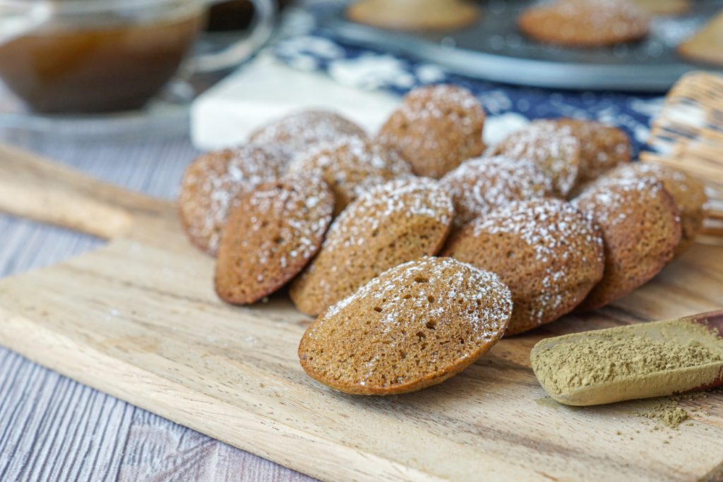 Hojicha Madeleines Thirteen Hojicha Madeleines on a wooden board next to hojicha powder.