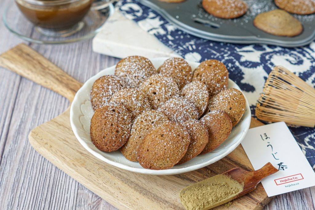 Hojicha Madeleines Madeleines on a white plate over a wooden board next to a small scoop of hojicha powder.