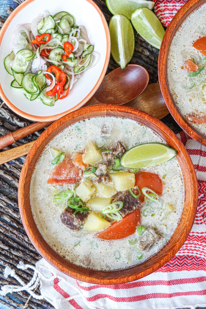 Aerial view of Soto Betawi (Betawi Beef and Coconut Soup) in two wooden bowls next to a red/white striped towel, two wooden spoons, lime wedges, and a cucumber pickle.