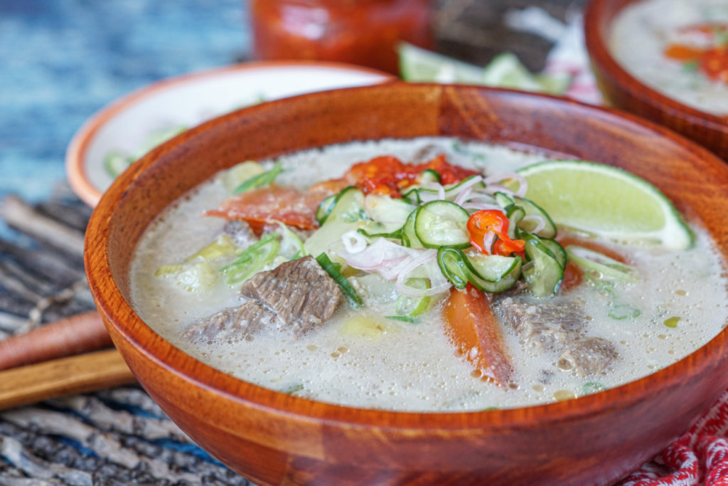 Close up of Soto Betawi (Betawi Beef and Coconut Soup) in a wooden bowl with lime wedges and a glass jar of Sambal Ulek in the background.