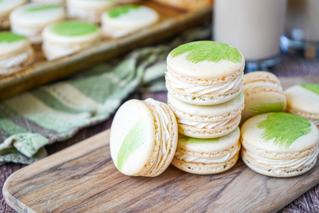 Irish Cream Macarons Stacked Irish Cream Macarons on a wooden board with more in the background on a baking sheet.