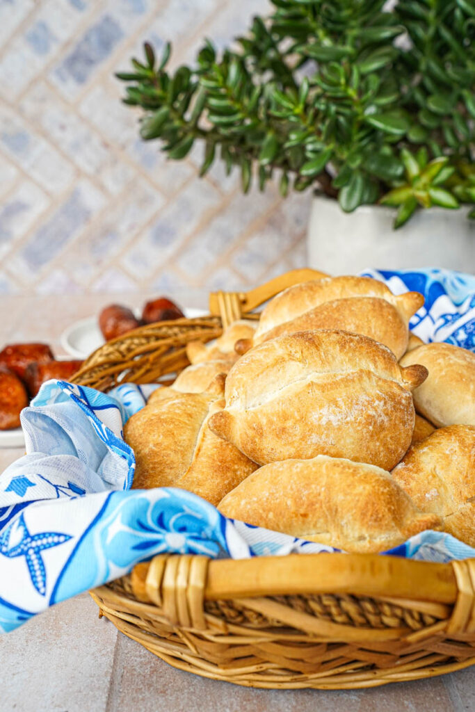 Side view of Papo Secos (Crusty Rolls) in a basket with two plates of sausages and a green plant in the background.