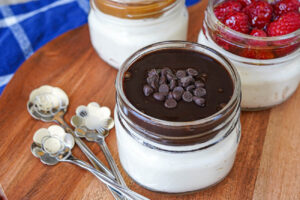 Three cheesecakes in jars topped with chocolate, raspberries, and dulce de leche.