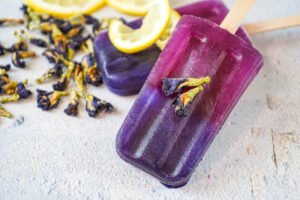 Two Butterfly Pea Flower Popsicles topped with butterfly pea flowers and lemon slices.