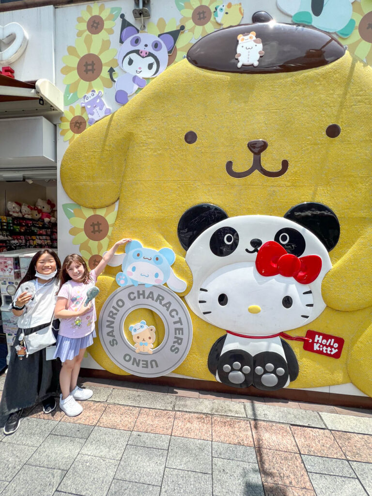 Two girls smiling in front of Sanrio store with Hello Kitty Panda and Pompompurin on side of building.