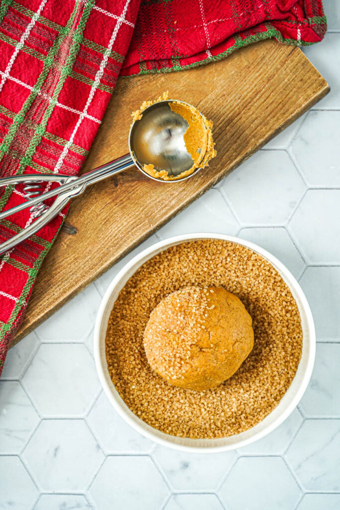 Aerial view of a ball of cookie dough in a white bowl of turbinado sugar next to an ice cream scoop.
