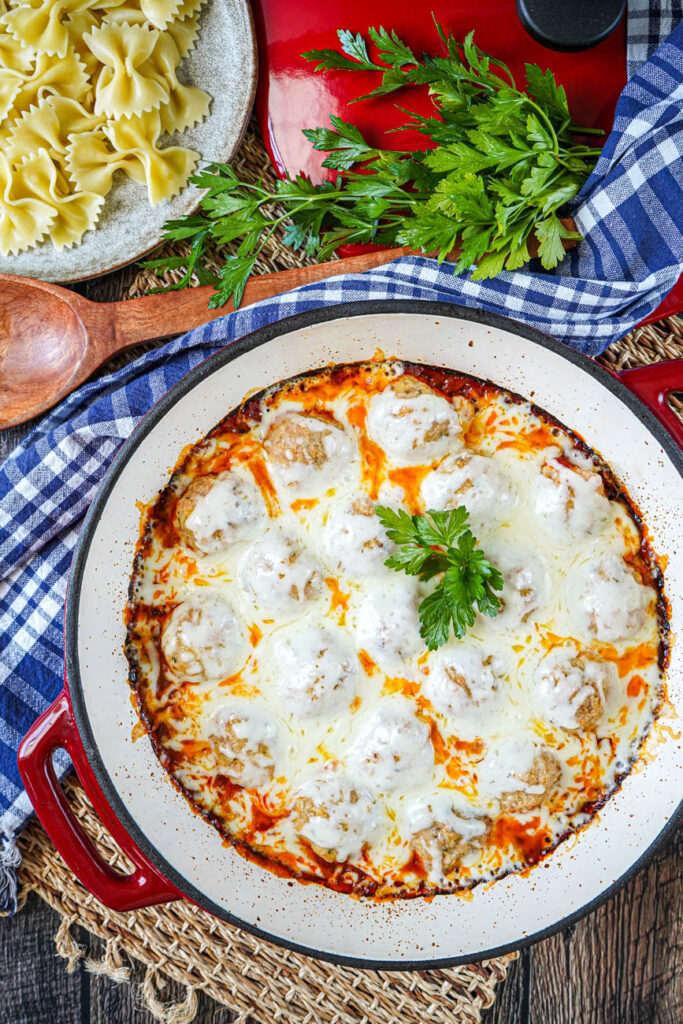 Aerial view of Effortless Chicken Meatball Marinara in a red pan next to a wooden spoon, parsley, and farfalle pasta.